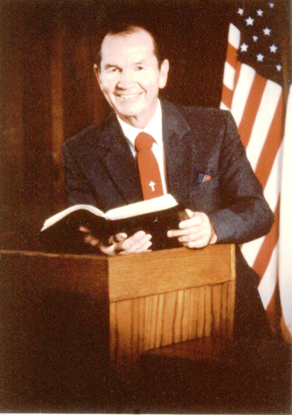 White man in suit and red tie with cross on it at lectern holding book with US flag behind