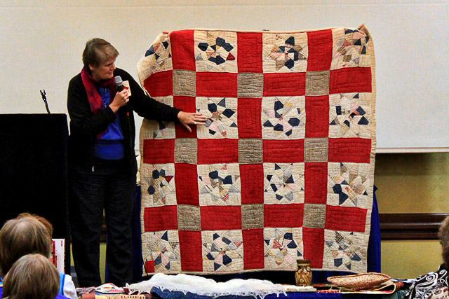 White woman talking into a microphone about a red and white quilt on stage
