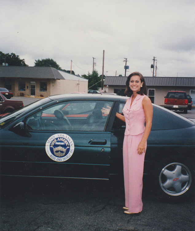 White woman in pink smiling with green car and single-story buildings in the background