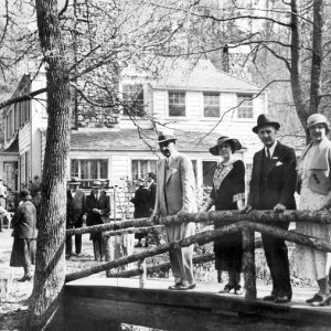 White men in suits and hats and women in hats and dresses pose on wooden bridge in front of two-story house and other people walking around