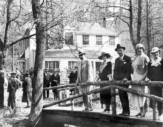 White men in suits and hats and women in hats and dresses pose on wooden bridge in front of two-story house and other people walking around