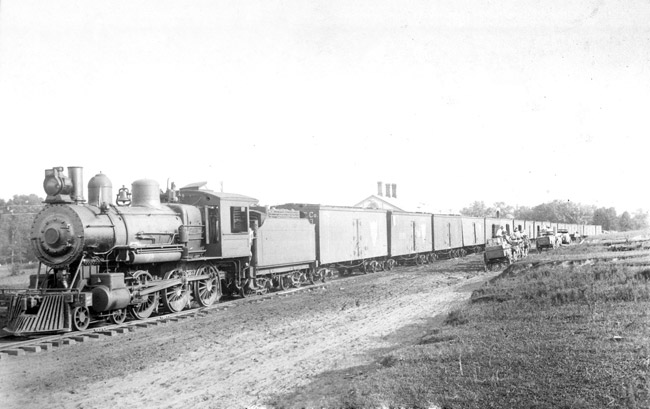 Steam locomotive with train cars on tracks and some trucks parked alongside
