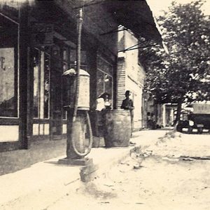 Patrons standing outside drug store building with gas pump on dirt road