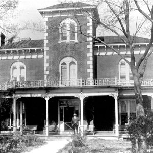 White man standing in front of a large brick house with an ornate porch and balcony