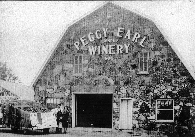 White man and white woman standing before large stone building "Peggy Earl Winery"