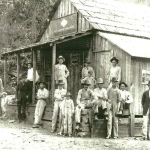 Group of white men standing and sitting on covered porch of single-story storefront