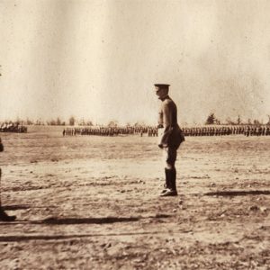 Three men in military uniform standing in a field with groups of soldiers standing in the background