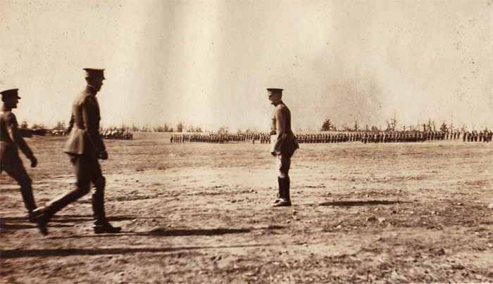 Three men in military uniform standing in a field with groups of soldiers standing in the background