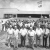 Group of old white men standing with mixed group of workers behind them with building in the background