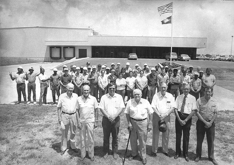 Group of old white men standing with mixed group of workers behind them with building in the background