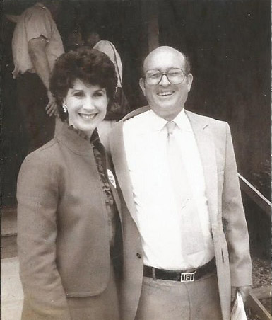 White woman smiling in suit standing with white man smiling in suit and glasses