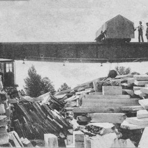 Piles of cut stone with three white men on quarrying equipment in the background