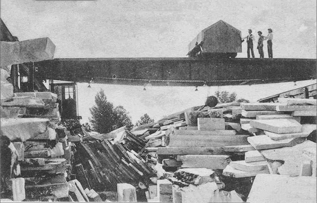 Piles of cut stone with three white men on quarrying equipment in the background
