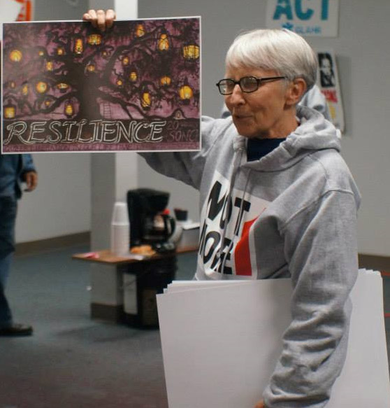 Older white woman with glasses in hooded sweater holding up a painting with the word "Resilience" on it