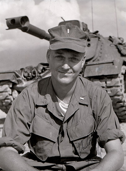 White man in military uniform with tank behind him