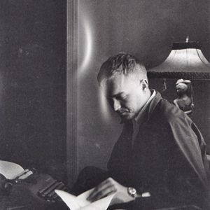 White man sitting at desk with typewriter and lamp behind him