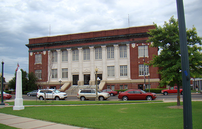 Multistory red-brick building with white columns and an obelisk monument on grass with parking lot and sidewalk