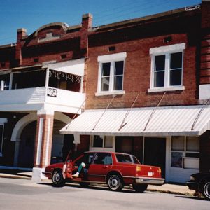 Two-story brick building with arched entrance way and covered balcony with red car in parking lot