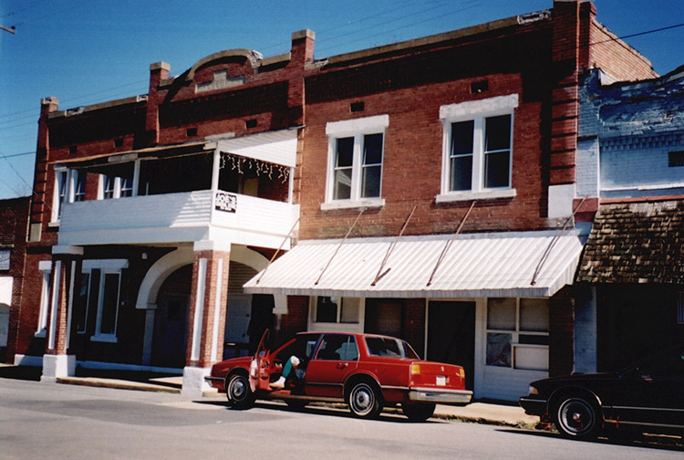 Two-story brick building with arched entrance way and covered balcony with red car in parking lot