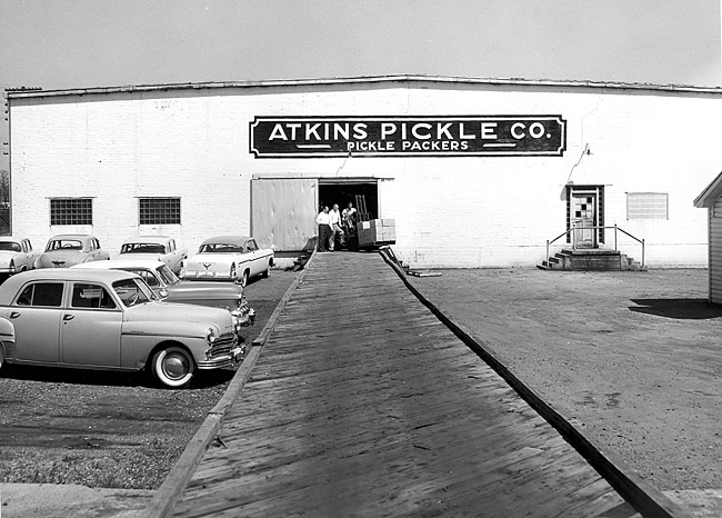 Group of white men and forklift exiting "Atkins Pickle Company" packing warehouse on parking lot with sign and cars
