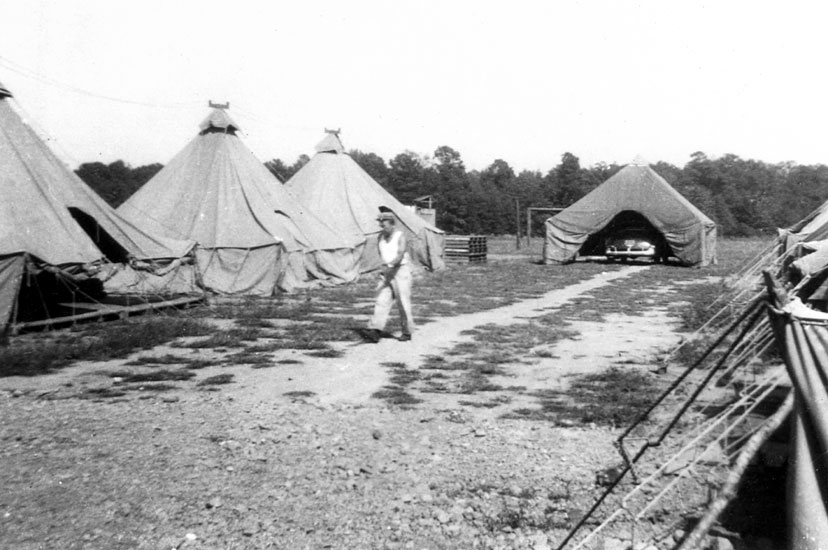 White man wearing German military helmet walking between rows of tents