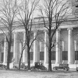 Large classical building with row of columns and windows with trees and parked cars on street