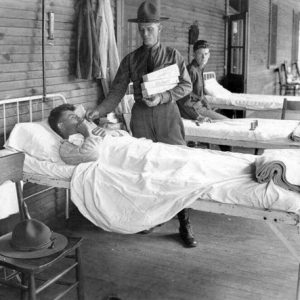 White man in uniform with hat handing out cigarettes to white patient in bed with white pillow and sheets and a hat on a chair in the foreground and another soldier sitting on a bed in the background