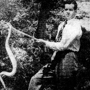 White man in shirt and tie holding up a snake with a stick