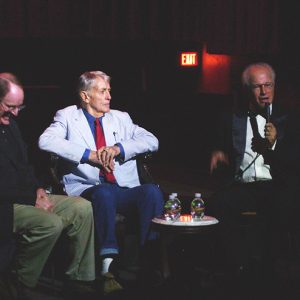 Three white men sitting in chairs on stage while one of them speaks into a microphone