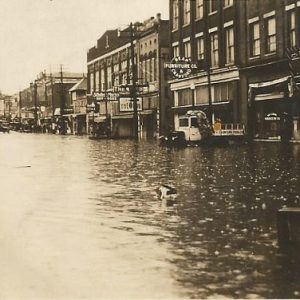 Flooded city street with brick storefronts and two-story building with clock tower in the distance