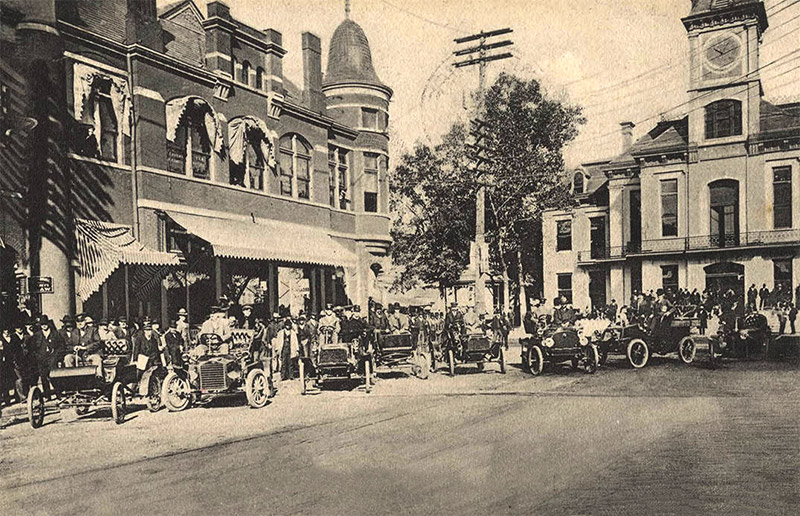 Crowd of men and their cars parked on street outside multistory buildings