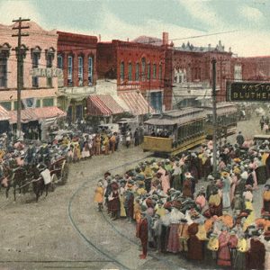 Crowded town street with multistory buildings on both sides and trolley cars running through its center