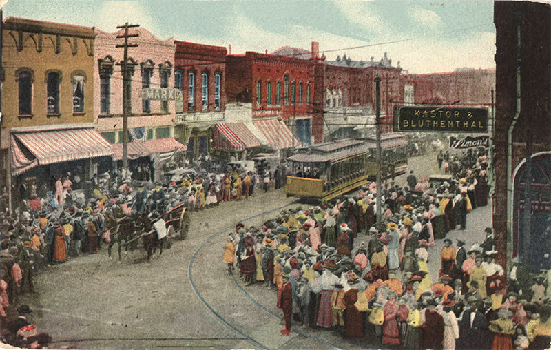Crowded town street with multistory buildings on both sides and trolley cars running through its center