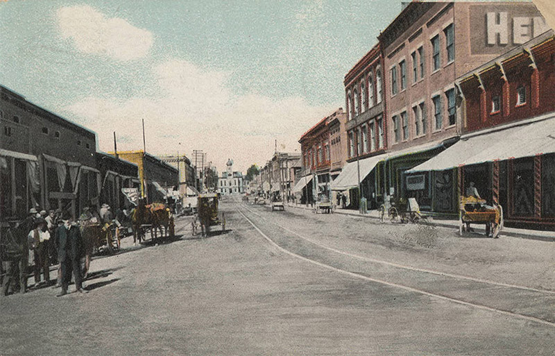 White men and horse drawn carriages on town street between rows of storefronts