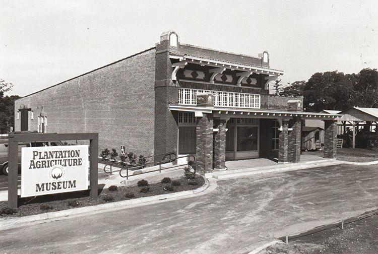 Two-story brick building with covered porch next to parking lot and sign "Plantation Agriculture Museum"