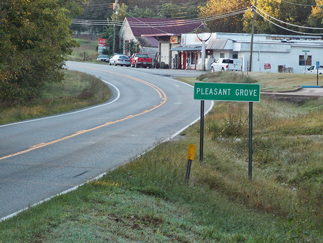 Single-story storefronts on two-lane street with "Pleasant Grove" sign in the foreground