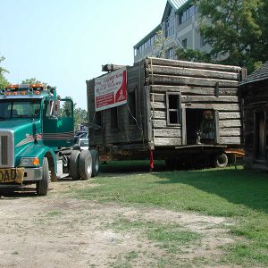 Semi-truck moving log house on trailer with multistory building in the background