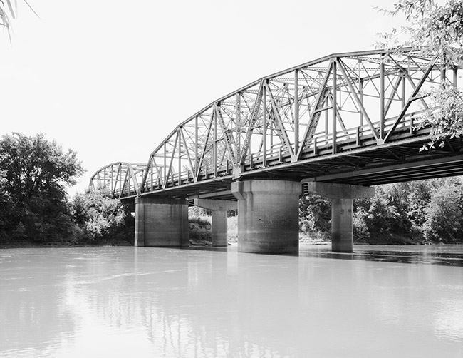 View from river bank of steel arch bridge with concrete supports in water
