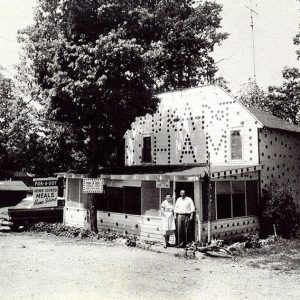 White man and woman standing outside two-story building with screened-in porch and polka-dot paint on dirt road