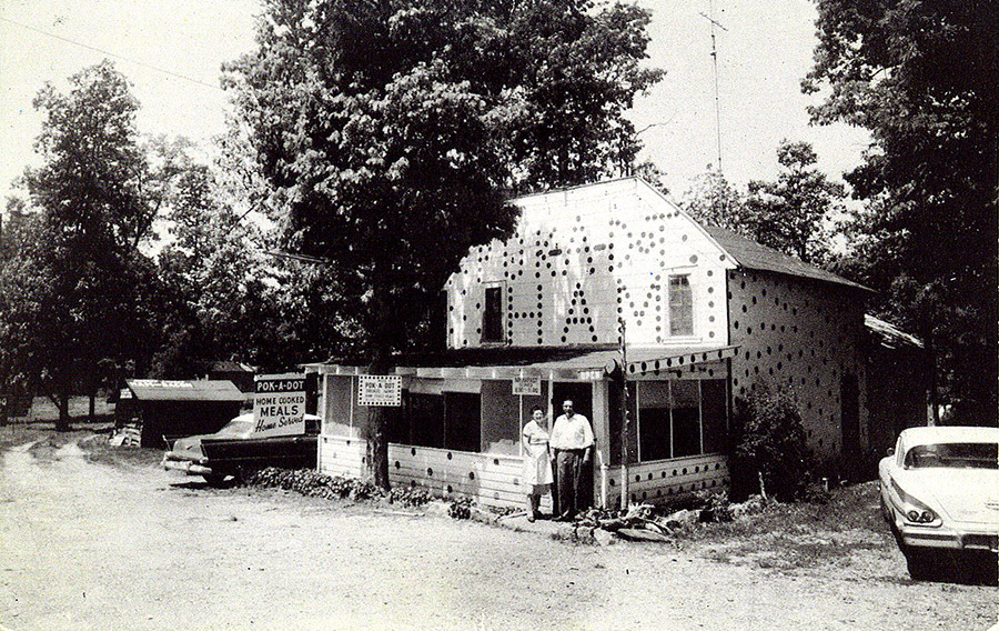 White man and woman standing outside two-story building with screened-in porch and polka-dot paint on dirt road