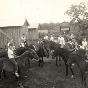 White man and boys on horseback with single-story cabins and barn behind them