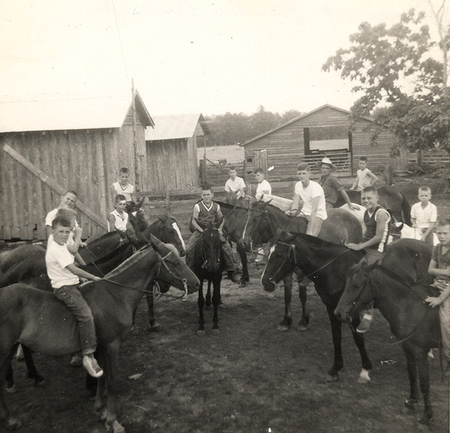White man and boys on horseback with single-story cabins and barn behind them