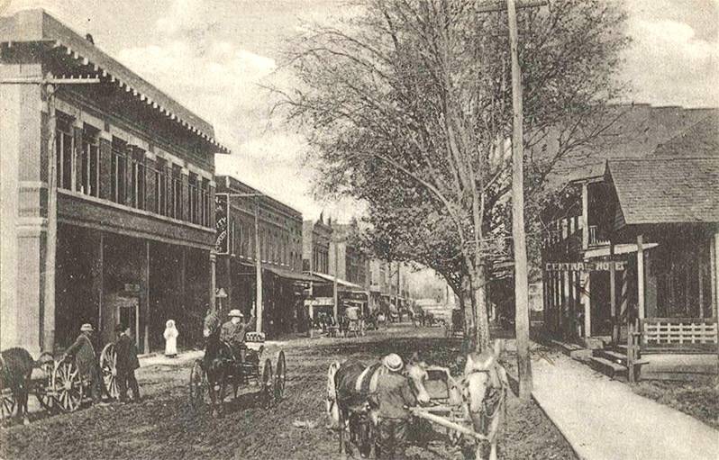 White men with horse drawn wagons on town street lined with buildings and trees