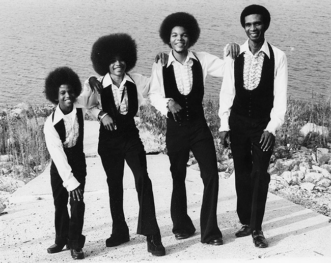 African-American man and three sons with afros and matching outfits of black pants and vests with white ruffled shirts standing together with body of water behind them