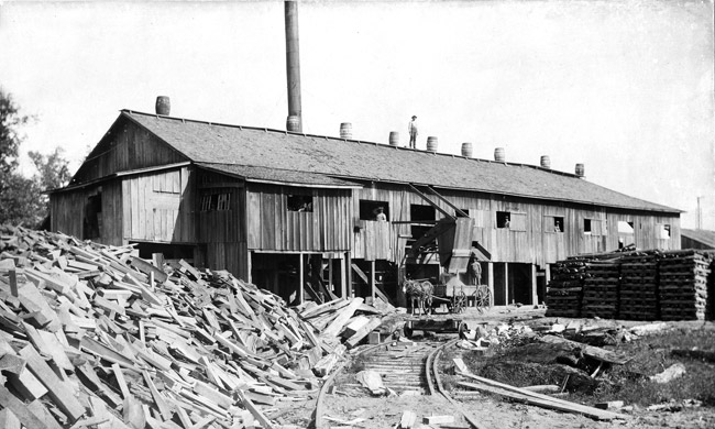 long wooden building with smoke stack next to railroad tracks and piles of lumber