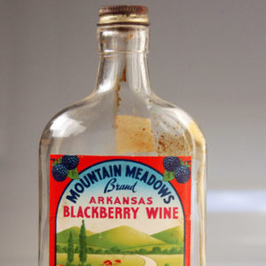 Empty screw top bottle with "Mountain Meadows brand Arkansas Blackberry Wine" label showing a bridge over a river and mountains on it