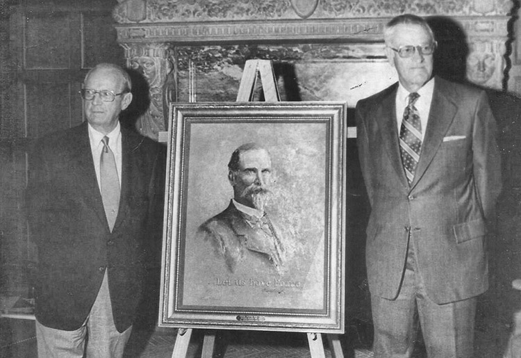 Pair of old white men with glasses in suit and tie standing at fireplace with framed painting of white man with beard in suit on easel in between them
