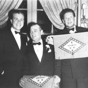 Three white men in tuxedos with one man holding an Arkansas flag standing in front of a set of french doors trimmed with heavy brocade curtains