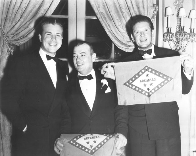 Three white men in tuxedos with one man holding an Arkansas flag standing in front of a set of french doors trimmed with heavy brocade curtains