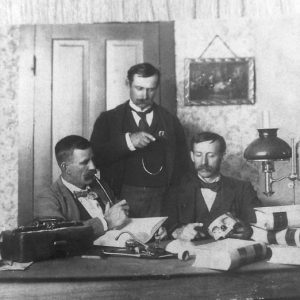 Three white men with mustaches and suits before a desk, one holding part of a human skull